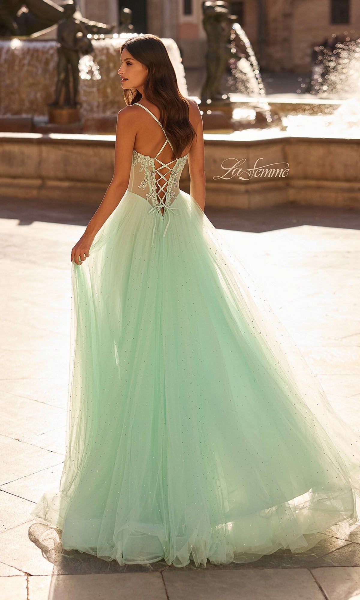 A woman wearing the La Femme 33860 long prom dress with a light green embroidered corset bodice and flowing tulle skirt stands outdoors by a fountain in daylight.